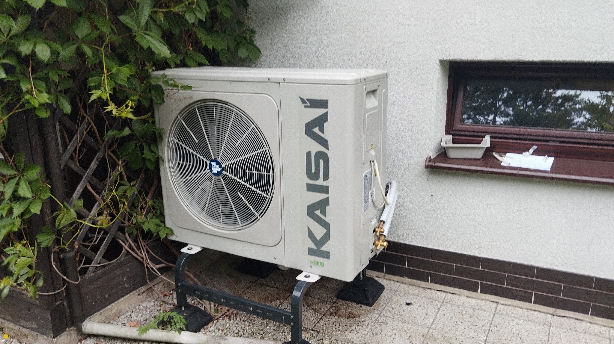 Zewnętrzna jednostka klimatyzacji Kaisai na metalowym stelażu, obok ściany z oknem i pnącą roślinnością, na tle jasnej elewacji i kostki brukowej.