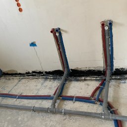 EkoHydro DAMIAN KACA - Wewnętrzne instalacje wodne i grzewcze w trakcie realizacji w Zielonej Górze, rury PEX w kolorach czerwonym i niebieskim, szare rury kanalizacyjne, mocowania do ściany, widoczne przewody elektryczne.