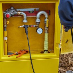 EkoHydro DAMIAN KACA - Żółta skrzynka gazowa z widocznymi rurami, reduktorem, manometrem i narzędziami, w tle ziemia.