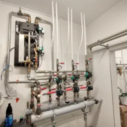 Rozbudowana instalacja grzewcza z licznymi rurami, zaworami i manometrami w kotłowni, widoczny panel sterowania i elementy zabezpieczeń przeciwpożarowych.