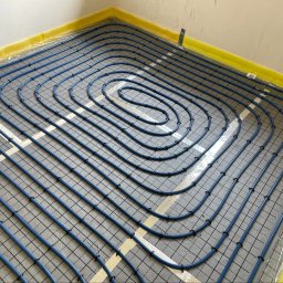 EkoHydro DAMIAN KACA - Ułożone niebieskie rury ogrzewania podłogowego w spiralnym wzorze na szarej macie z siatką, zabezpieczone klipsami, z żółtą izolacją brzegową w narożniku pomieszczenia.