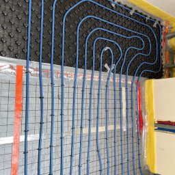 EkoHydro DAMIAN KACA - Instalacja niebieskich rur ogrzewania podłogowego na czarnej macie z wypustkami i szarej siatce, z widoczną żółtą izolacją przy ścianie w Zielonej Górze.