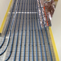 EkoHydro DAMIAN KACA - Niebieskie rury ogrzewania podłogowego ułożone równolegle na szarej macie izolacyjnej z siatką, przymocowane czarnymi klipsami, z żółtą izolacją brzegową.