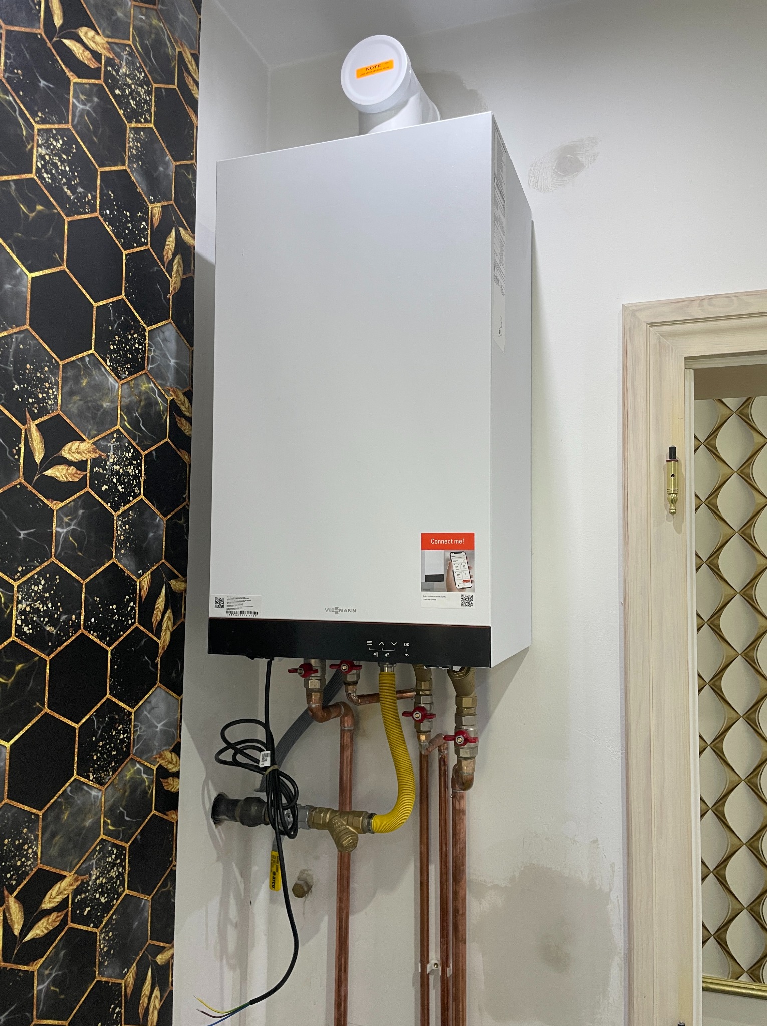 Nowoczesny, biały piec gazowy Viessmann zamontowany na ścianie z dekoracyjną tapetą w geometryczne wzory, widoczne miedziane rury i żółty przewód gazowy, obok framuga drzwi z ozdobnym wypełnieniem.