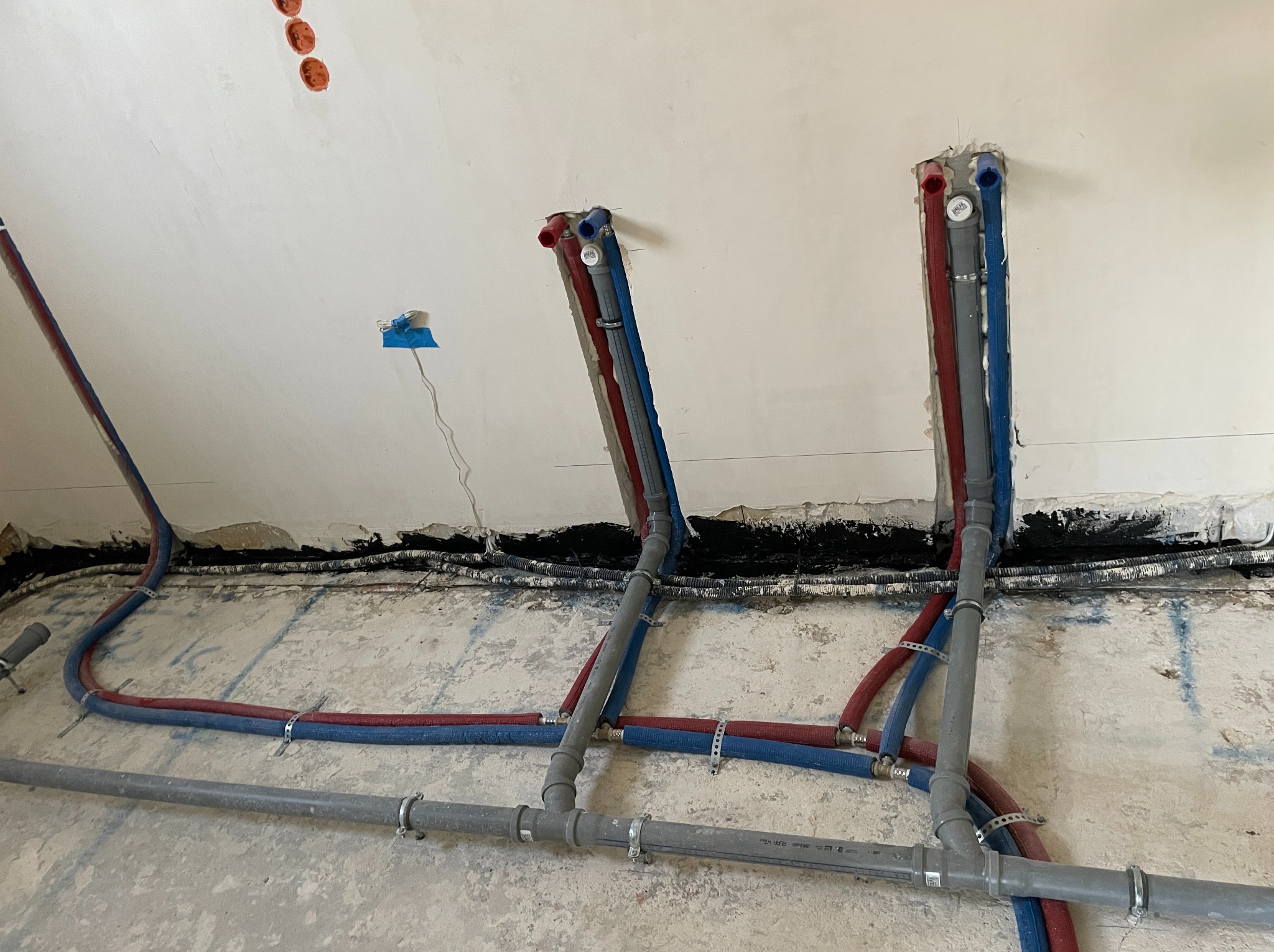 Wewnętrzne instalacje wodne i grzewcze w trakcie realizacji w Zielonej Górze, rury PEX w kolorach czerwonym i niebieskim, szare rury kanalizacyjne, mocowania do ściany, widoczne przewody elektryczne.