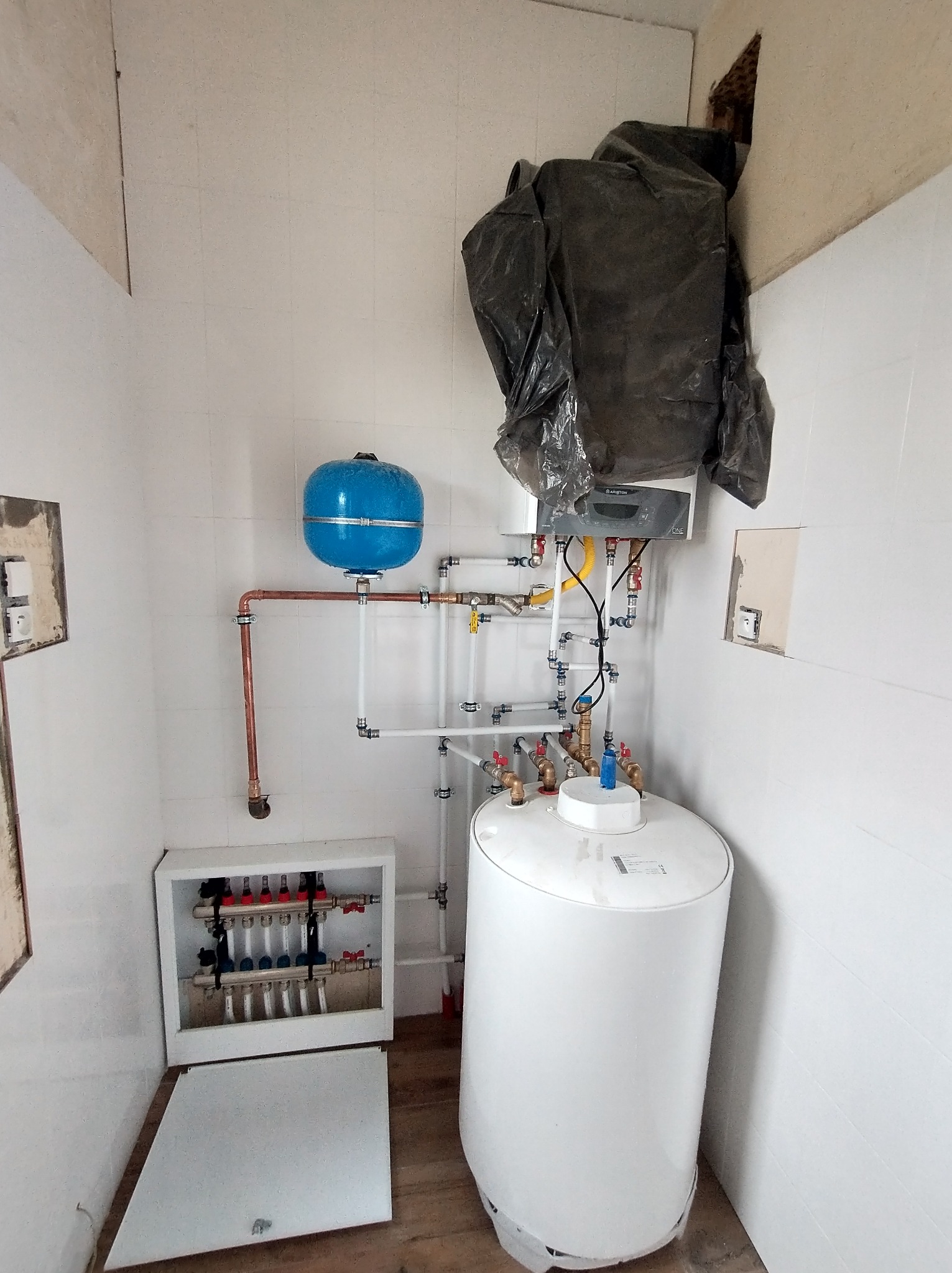 Wnętrze kotłowni z zamontowanym piecem gazowym Ariston przykrytym folią, zbiornikiem wody, niebieskim naczyniem przeponowym, rozdzielaczem grzewczym w szafce z otwartymi drzwiczkami oraz instalacją...