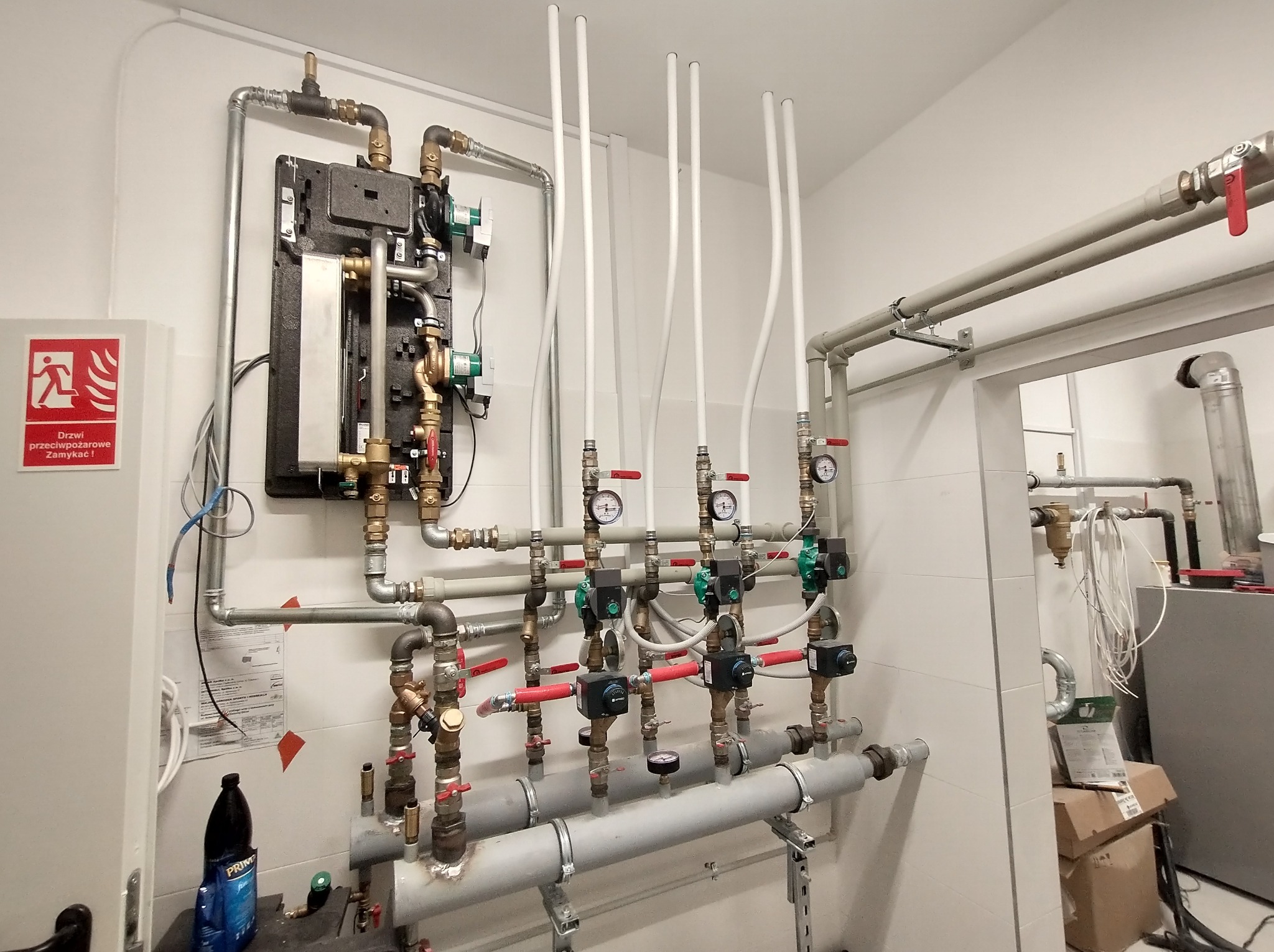 Rozbudowana instalacja grzewcza z licznymi rurami, zaworami i manometrami w kotłowni, widoczny panel sterowania i elementy zabezpieczeń przeciwpożarowych.