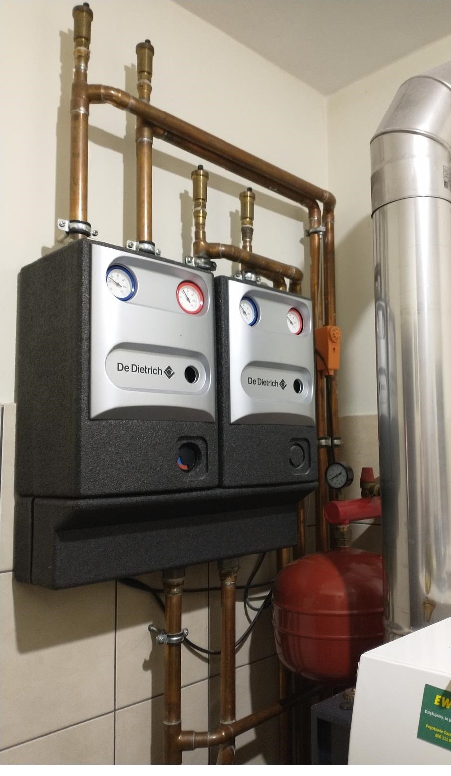 Dwa kotły gazowe marki De Dietrich zamontowane na ścianie z widocznymi rurami miedzianymi, manometrami i zaworami, z czerwonym zbiornikiem wyrównawczym w tle.