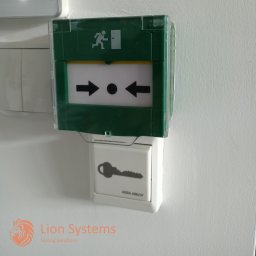 PPHU LionSystems Dmytro Stasiuk - Przycisk alarmowy z piktogramem biegnącej osoby i strzałkami wskazującymi kierunek, zamontowany na ścianie obok włącznika z symbolem klucza.