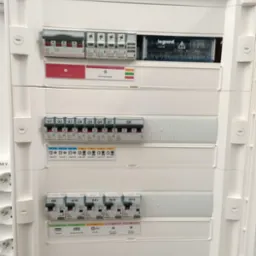 Biała skrzynka elektryczna z bezpiecznikami i wyłącznikami firmy Legrand, widoczne oznaczenia Q1-Q13 i diody sygnalizacyjne.