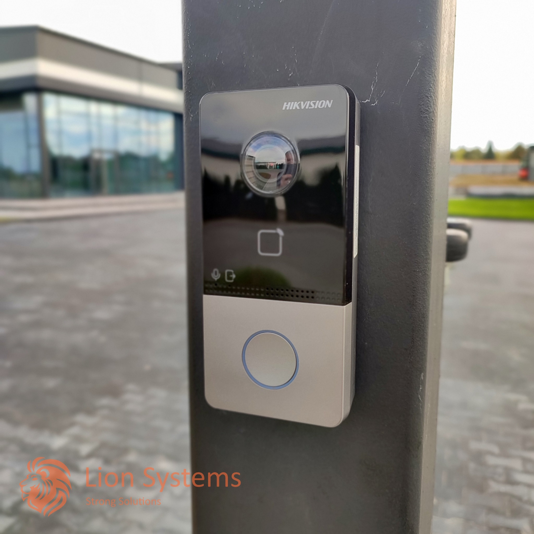 Zewnętrzny panel domofonowy Hikvision z kamerą na słupku ogrodzenia, widoczny budynek w tle.