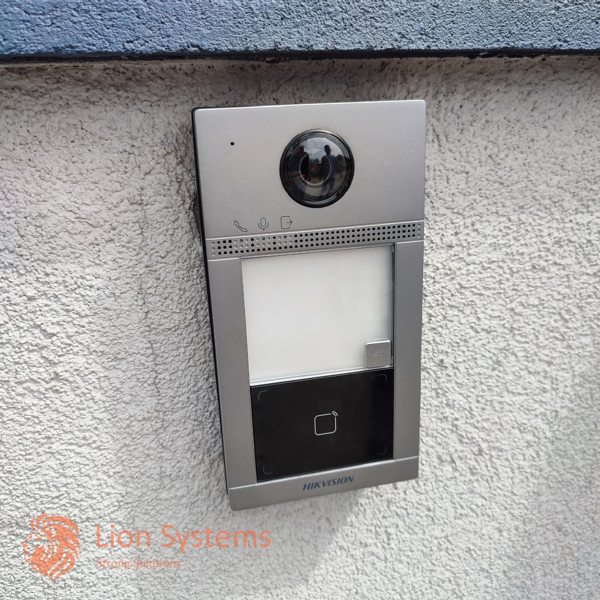 Zewnętrzny panel domofonowy Hikvision z kamerą i czytnikiem kart zbliżeniowych, zamontowany na białej, szorstkiej ścianie z niebieskim akcentem u góry.