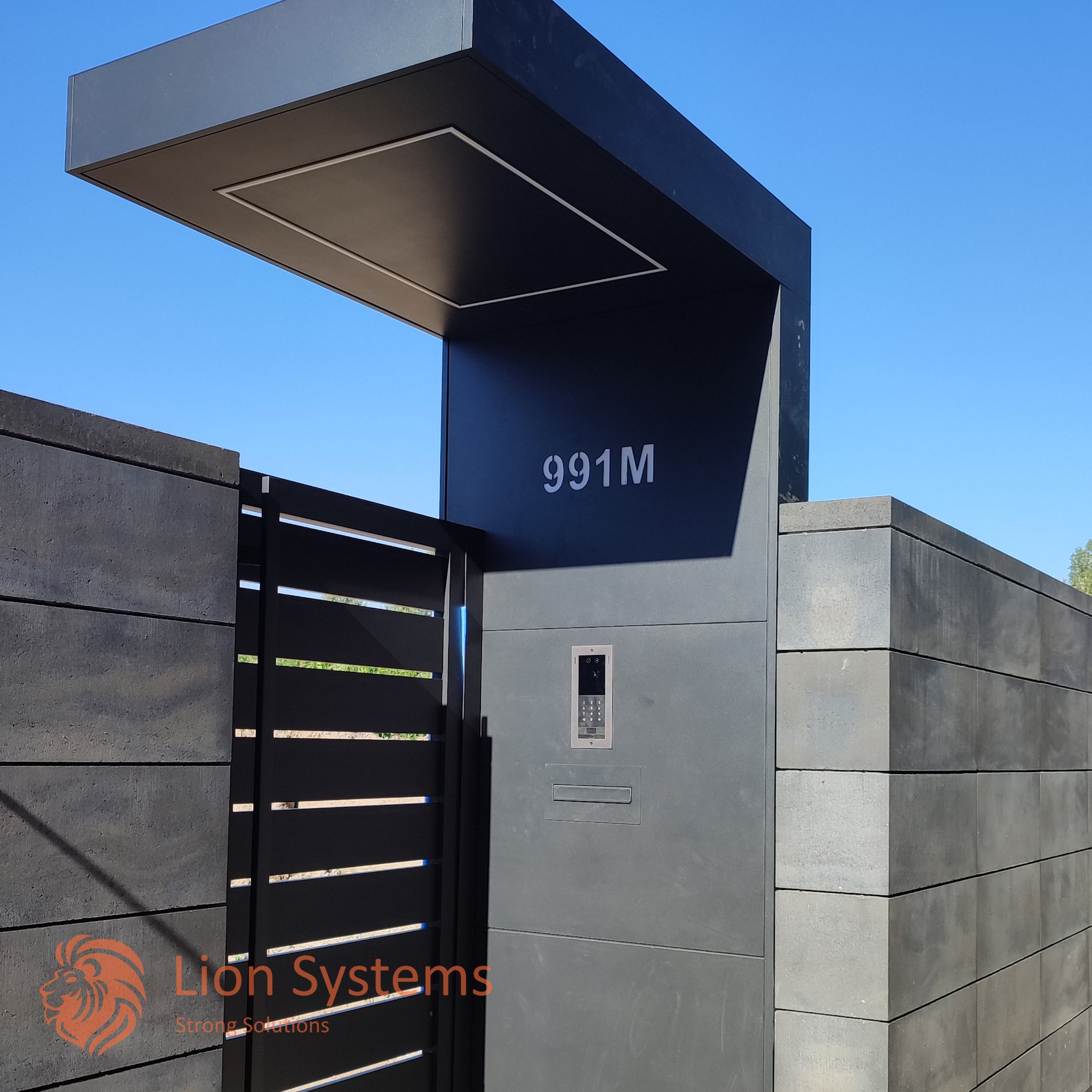 Nowoczesna brama wjazdowa z panelem domofonowym, numerem domu '991M' i zadaszeniem, wykonana z szarych elementów betonowych i czarnych paneli poziomych.