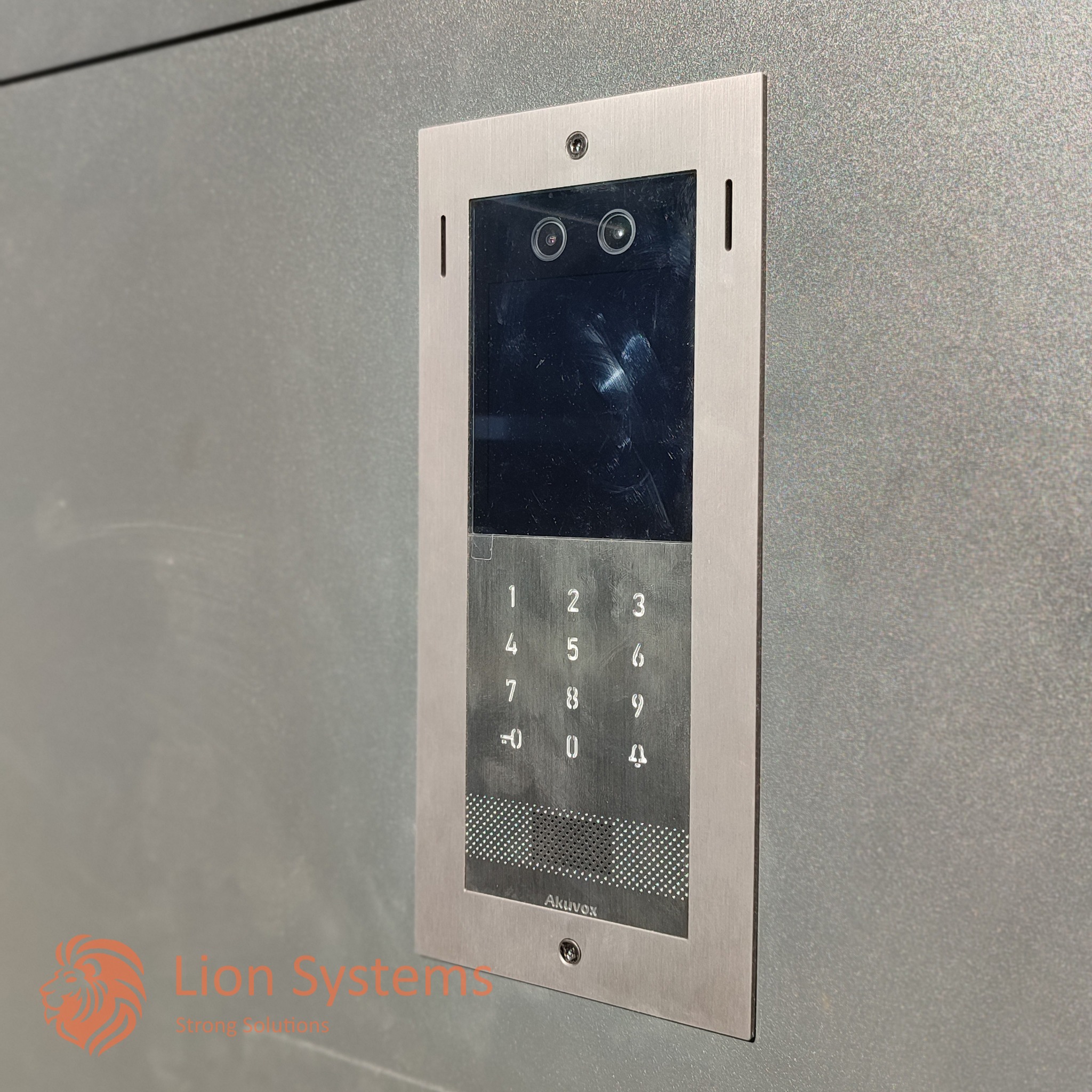 Metalowy panel domofonu z kamerą i klawiaturą numeryczną, zamontowany na szarej ścianie. Widoczne dwie soczewki obiektywu kamery i logo Akuvox na dole.