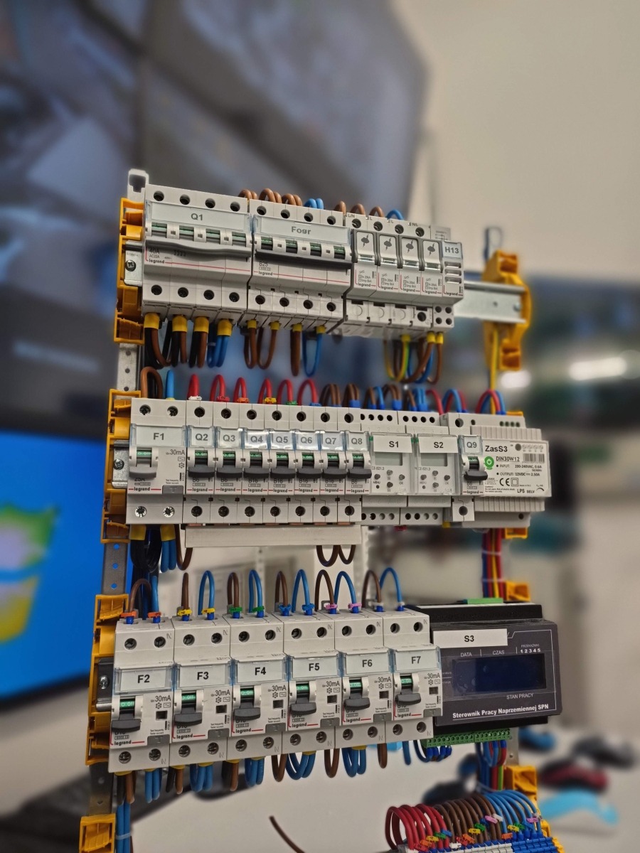 Rozdzielnica elektryczna z zamontowanymi bezpiecznikami i przewodami o różnych kolorach, widoczne oznaczenia F1-F7, Q1-Q9, S1-S3 oraz sterownik pracy naprzemiennej SPN.