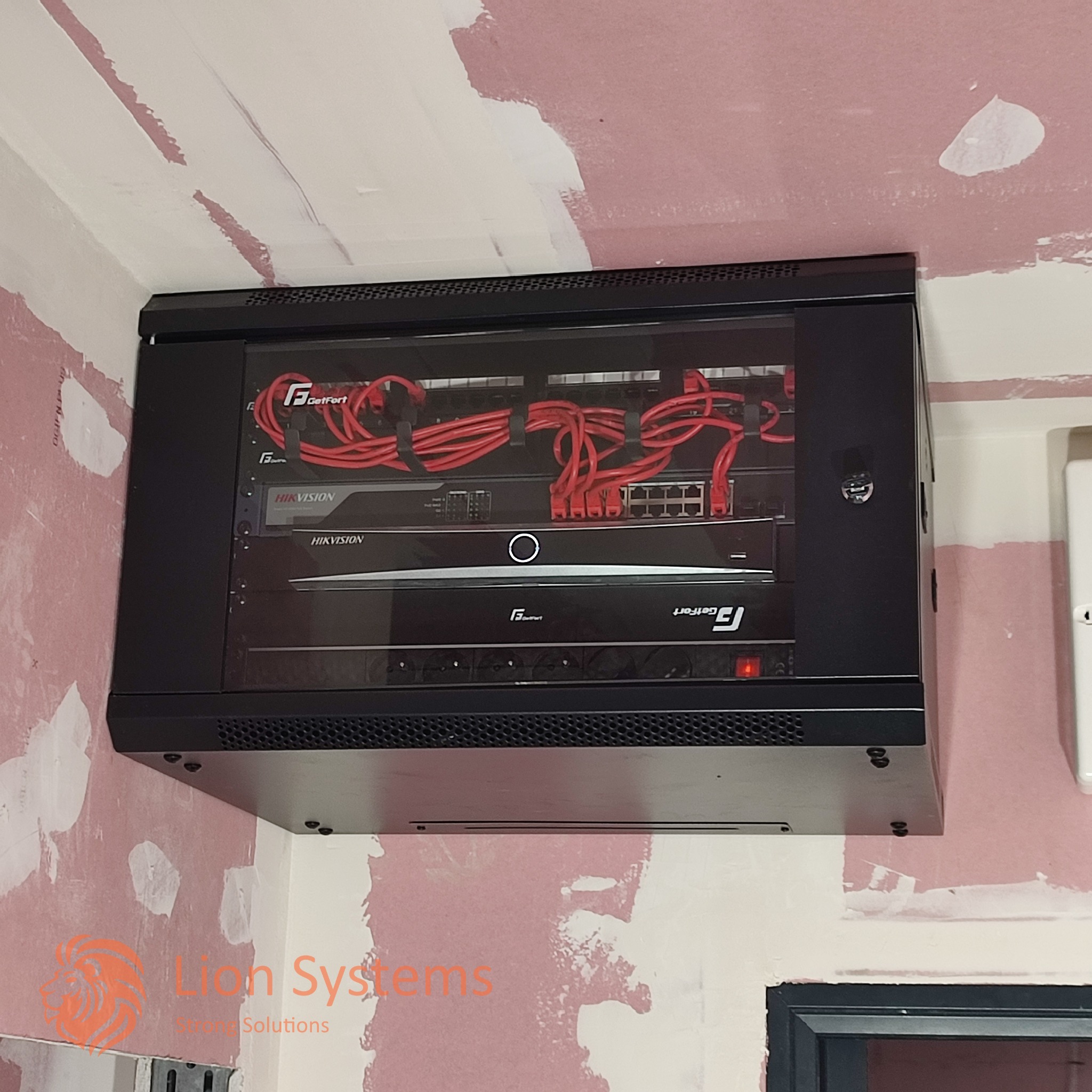 Czarna szafa rack z zainstalowanymi urządzeniami Hikvision i GetFort, okablowanie w kolorze czerwonym, zamontowana na ścianie w trakcie remontu.