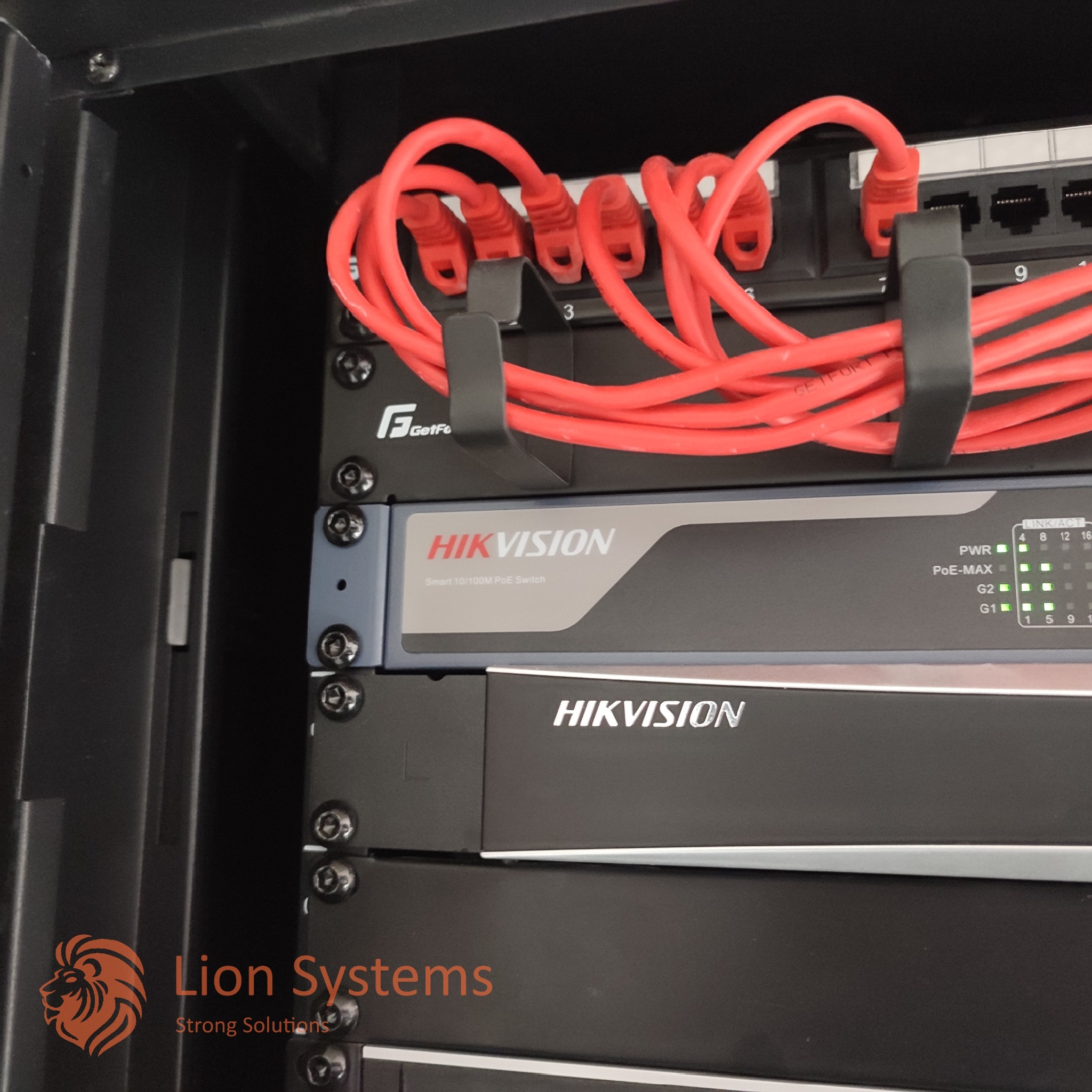 Zamknięta szafa rack z zainstalowanym switchem Hikvision Smart 10/100M PoE, podłączonymi czerwonymi kablami Ethernet oraz widocznymi diodami sygnalizującymi pracę urządzenia.