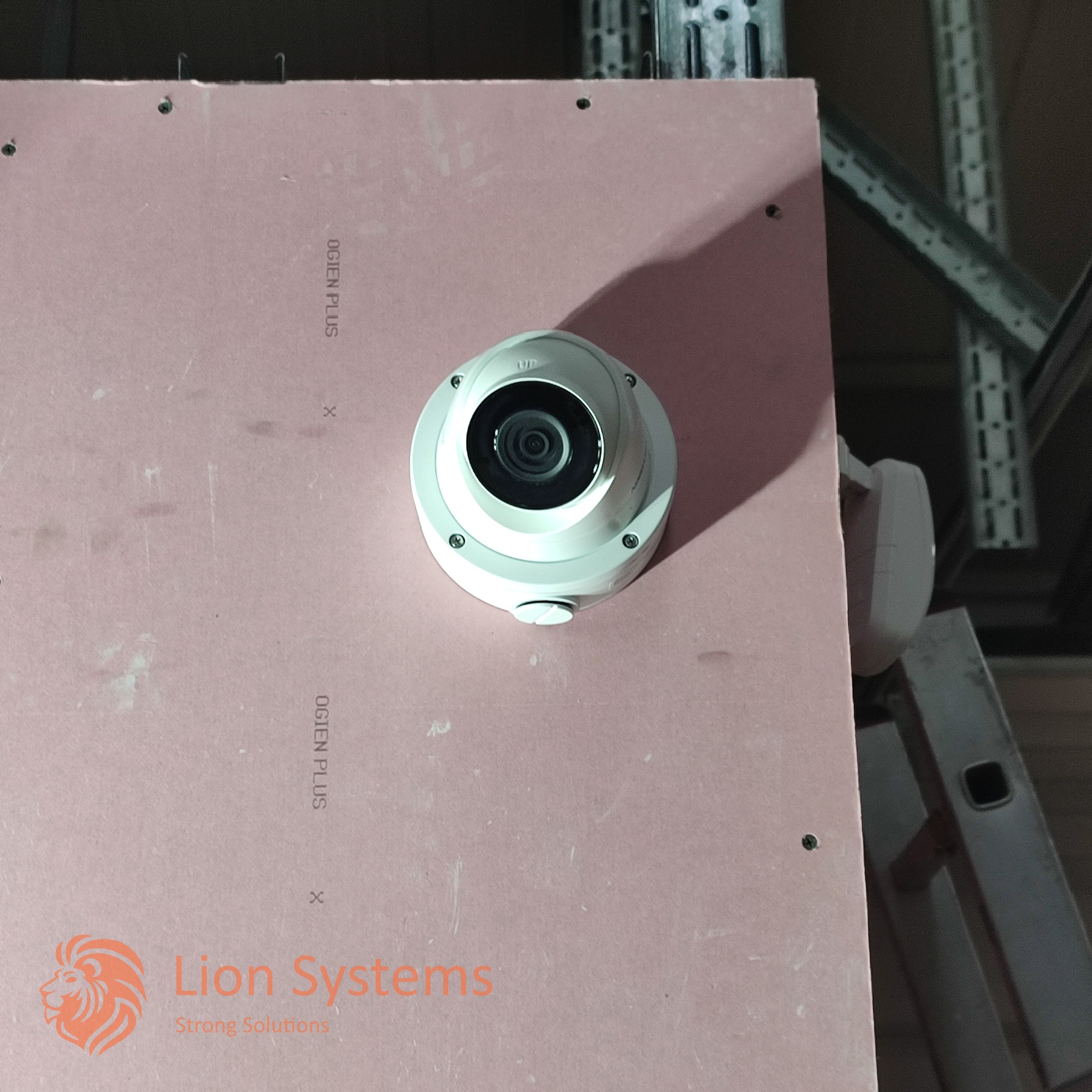 Kopułkowa kamera monitoringu zamontowana na różowej płycie gipsowej z widocznym logo firmy Lion Systems w rogu, w tle metalowa konstrukcja i fragment drabiny