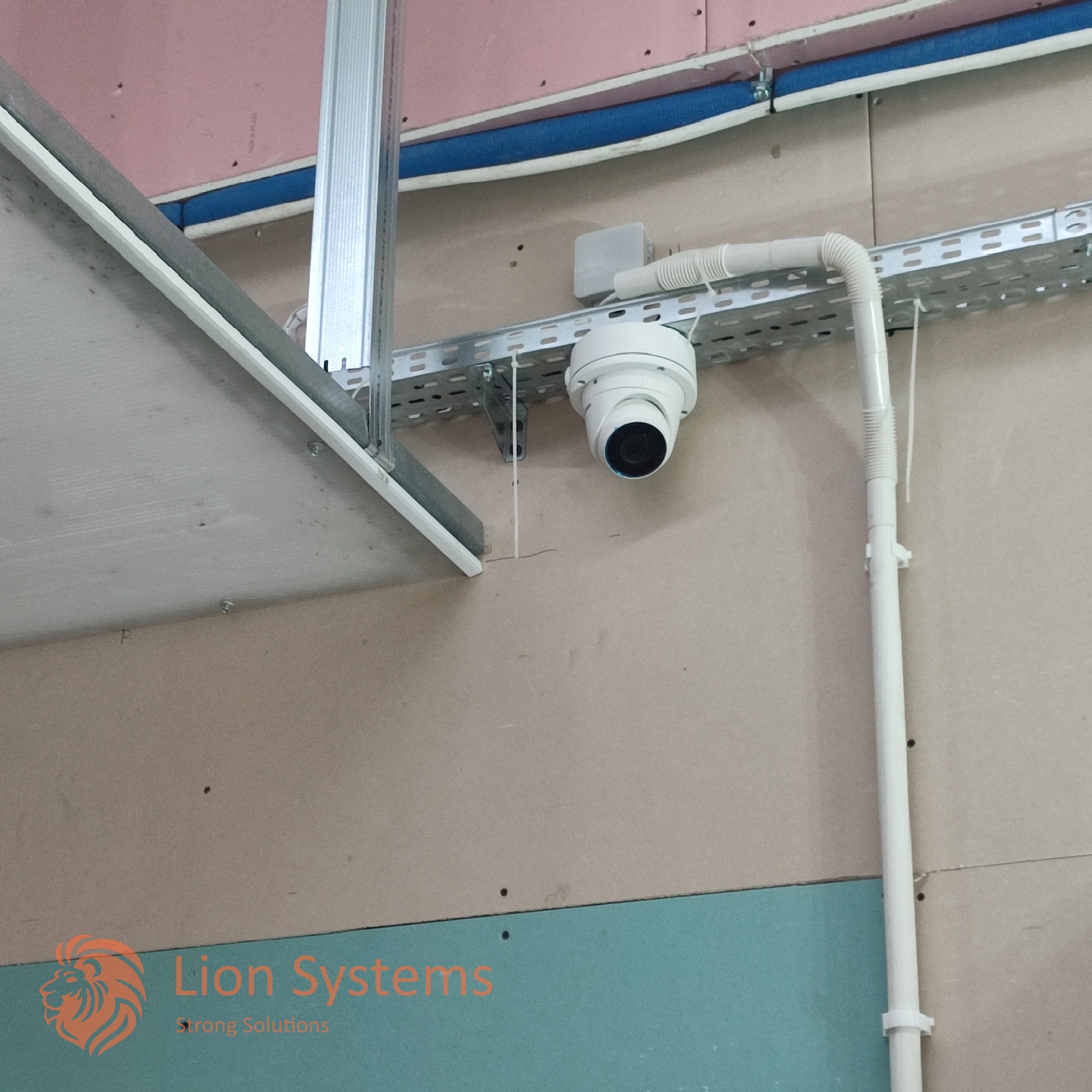 Kopułkowa kamera monitoringu zamontowana na metalowej szynie pod sufitem podwieszanym w trakcie montażu, widoczne przewody i elementy konstrukcyjne.