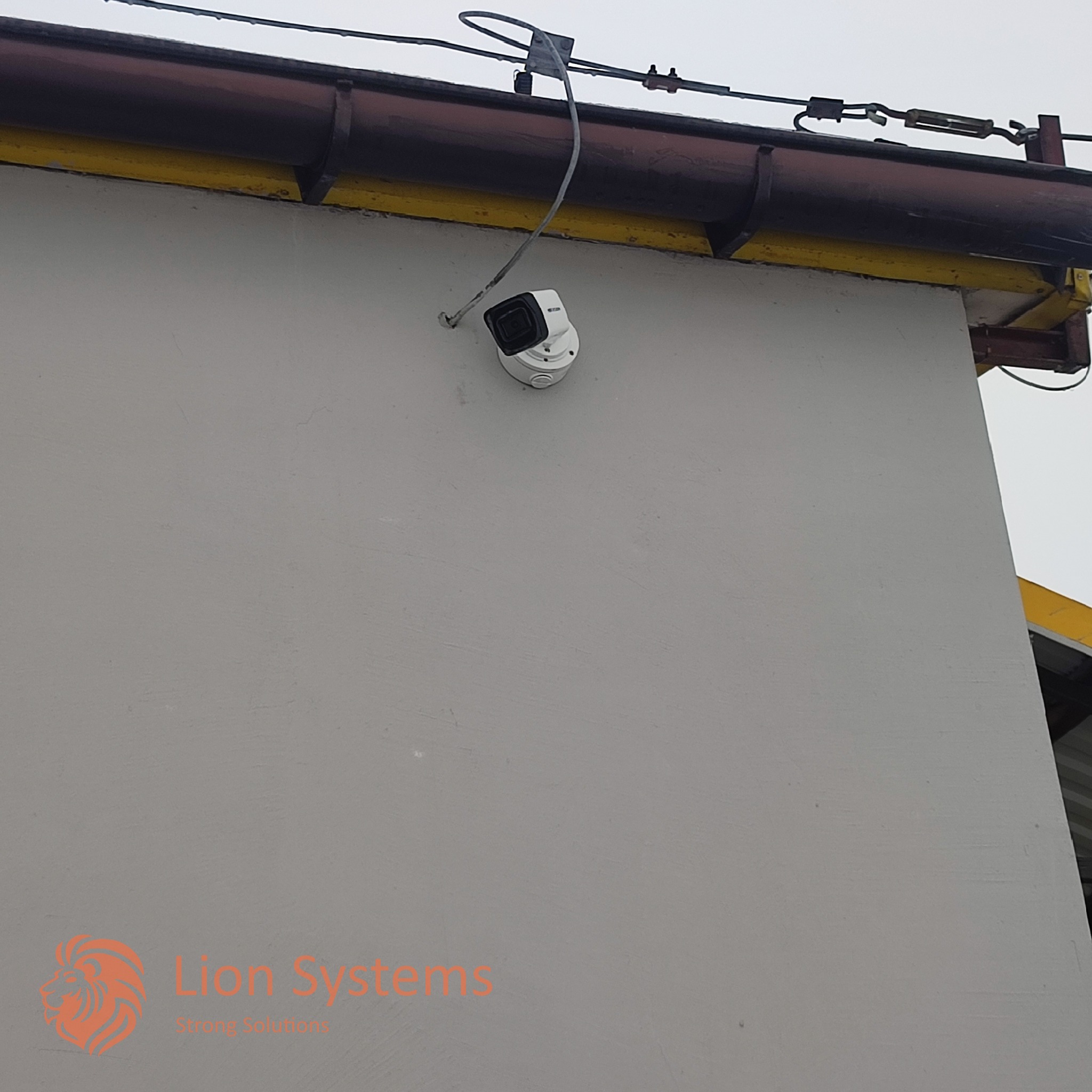 Zewnętrzna kamera monitoringu w obudowie typu bullet zamontowana na szarej ścianie budynku, widoczne przewody i elementy mocujące pod dachem z brązową rynną.