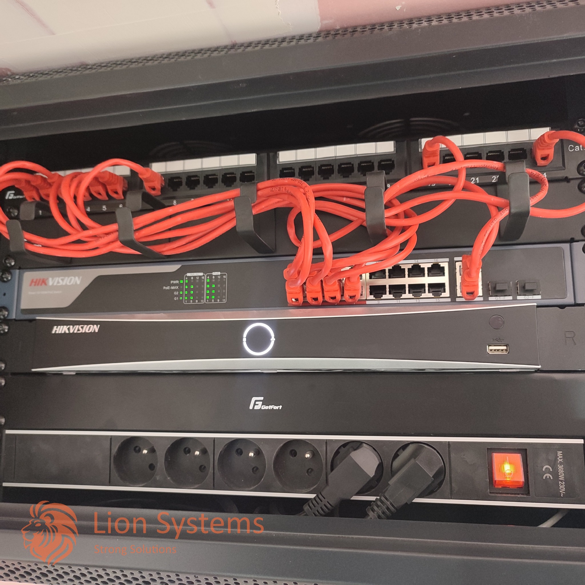 Szafa rack z zainstalowanymi urządzeniami sieciowymi Hikvision i GetFort, patch panelem i listwą zasilającą z włączonym czerwonym przełącznikiem, okablowanie w kolorze czerwonym.