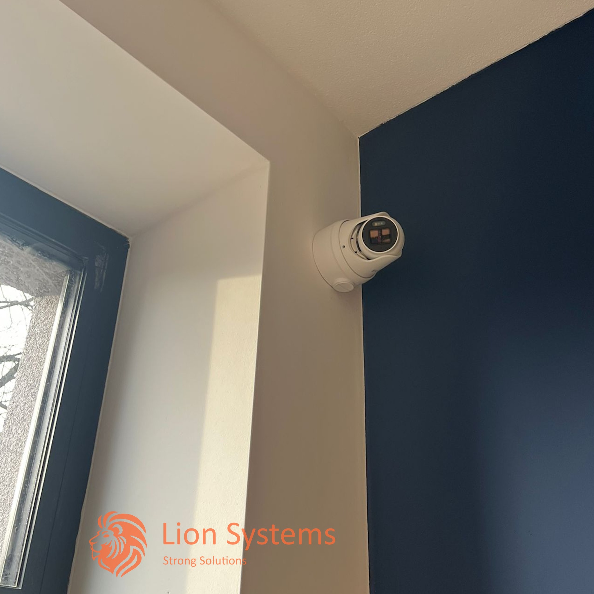 Kamera monitoringu zamontowana w rogu pomieszczenia, widoczne fragmenty okna i ściany pomalowanej na granatowo.