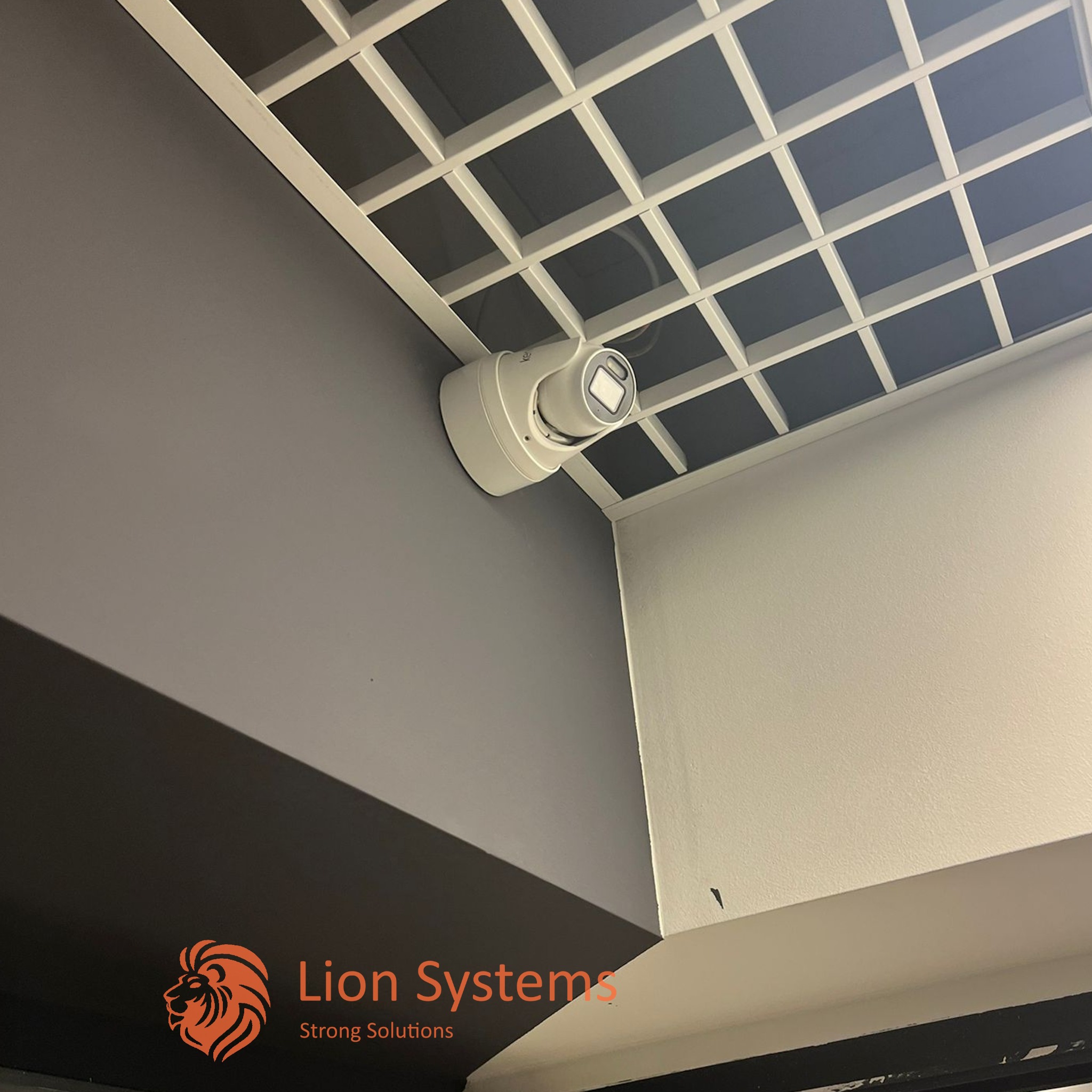 Kamera monitoringu kopułkowa zamontowana pod sufitem rastrowym w pomieszczeniu biurowym, widoczny fragment ściany szarej i białej, logo firmy w rogu kadru.