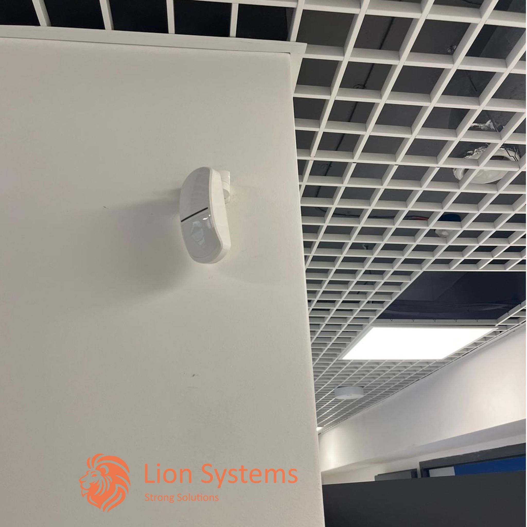 Biały czujnik ruchu zamontowany na ścianie w biurze w Rzeszowie, widoczny sufit rastrowy z oświetleniem.