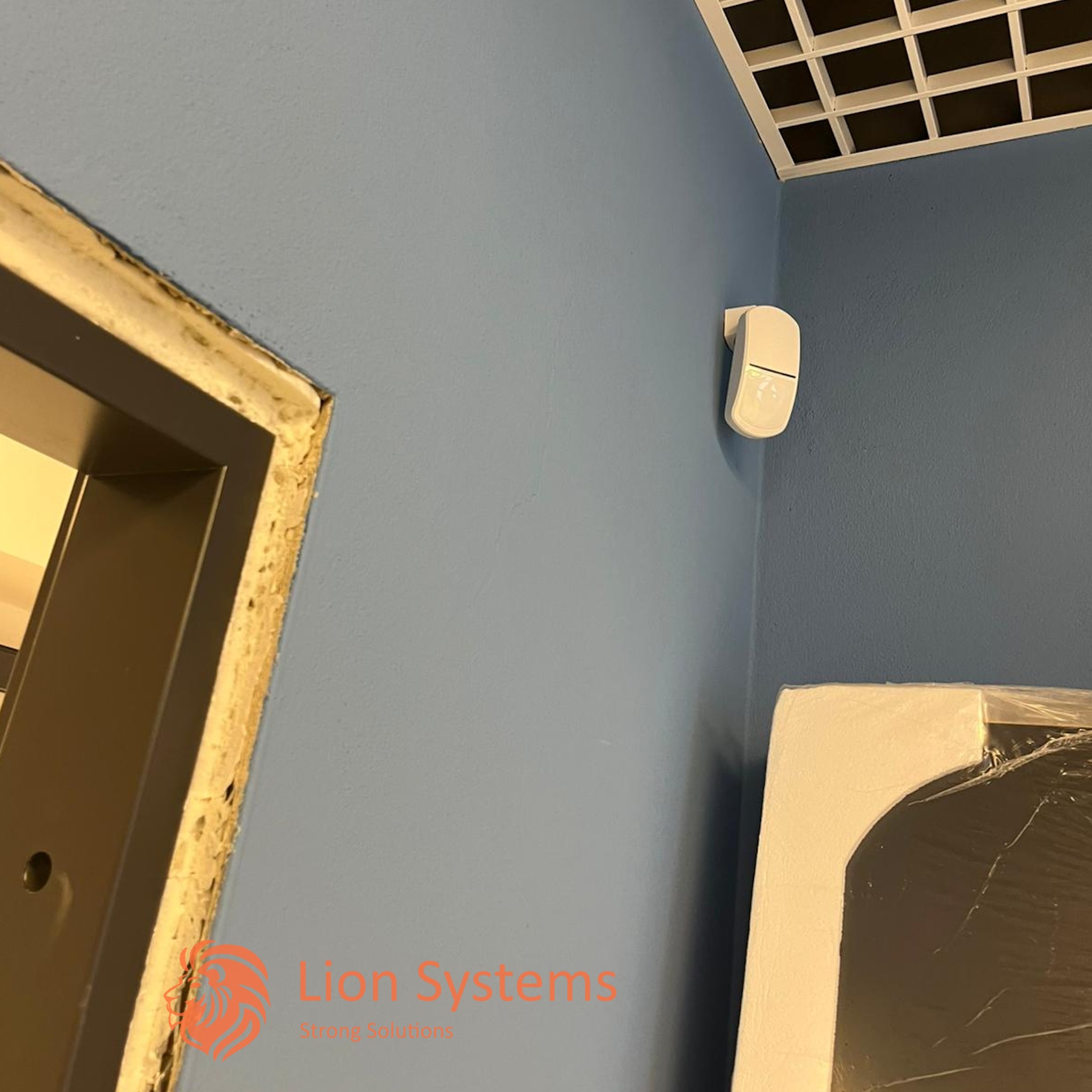 Biały czujnik ruchu zamontowany na niebieskiej ścianie w rogu pomieszczenia, widoczny fragment sufitu kasetonowego i ościeżnicy drzwi.