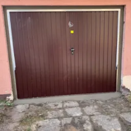 Brązowa, metalowa brama garażowa z pionowymi panelami, świeżo osadzona w ościeżnicy, widoczne ślady cementu i niedoskonałości tynku na ścianie.