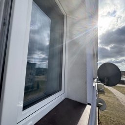 Robud Robert Małkiewicz - Białe okno z widokiem na krajobraz miejski i anteny satelitarne, oświetlone promieniami słonecznymi.