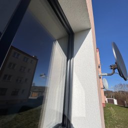Robud Robert Małkiewicz - Szare okno z widocznym w szybie odbiciem budynku i nieba, obok elewacja budynku z anteną satelitarną na tle błękitnego nieba.