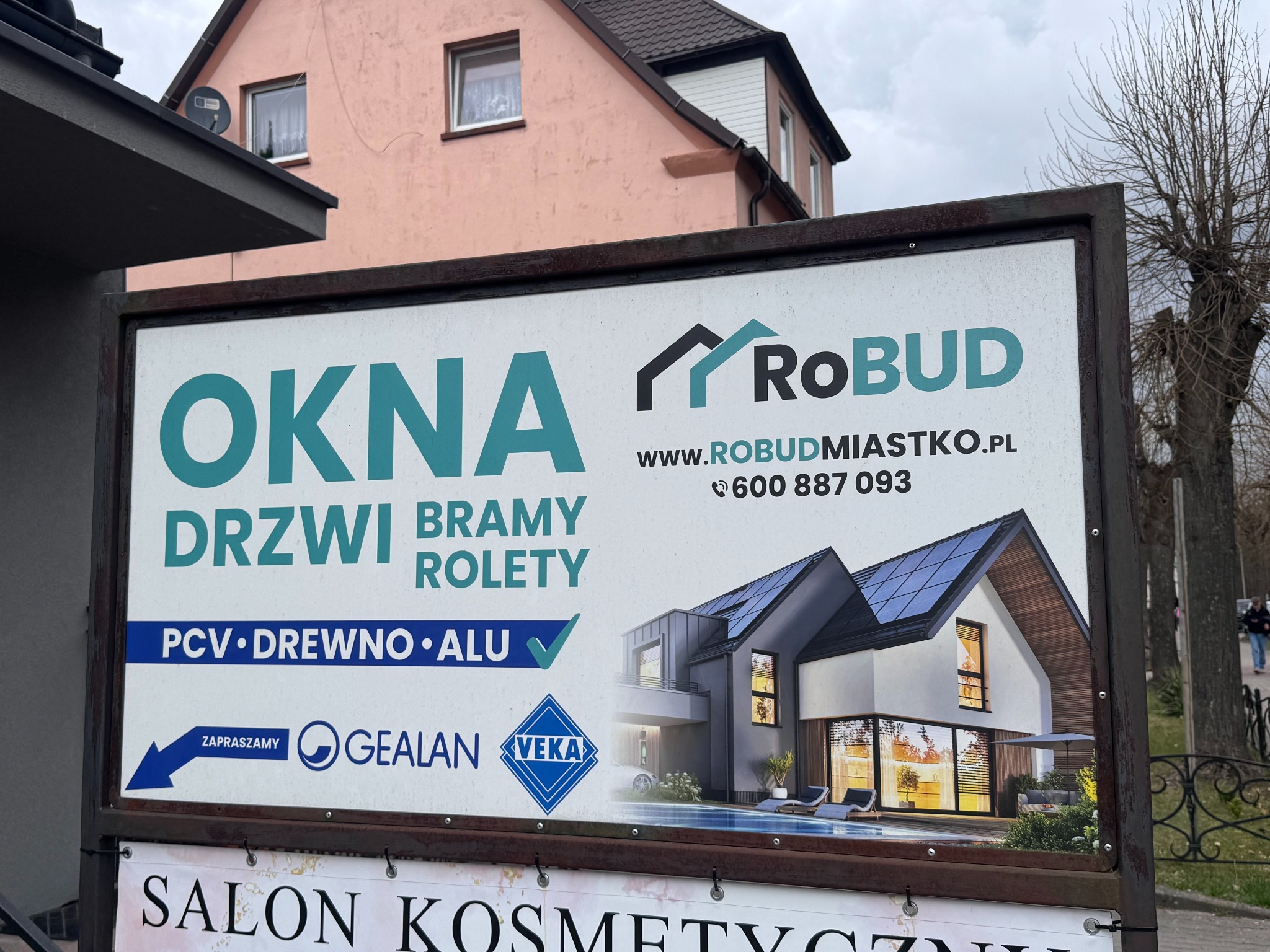 Billboard firmy Robud z Miastka reklamujący okna, drzwi, bramy i rolety z PCV, drewna i aluminium, z wizualizacją nowoczesnego domu z basenem.