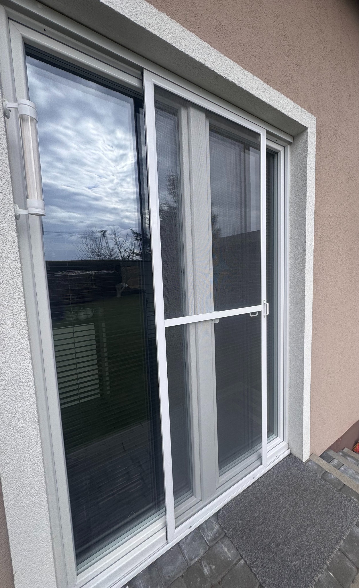 Przesuwne okno z moskitierą, widok z zewnątrz, szara wycieraczka przed oknem, pochmurne niebo odbija się w szybie.