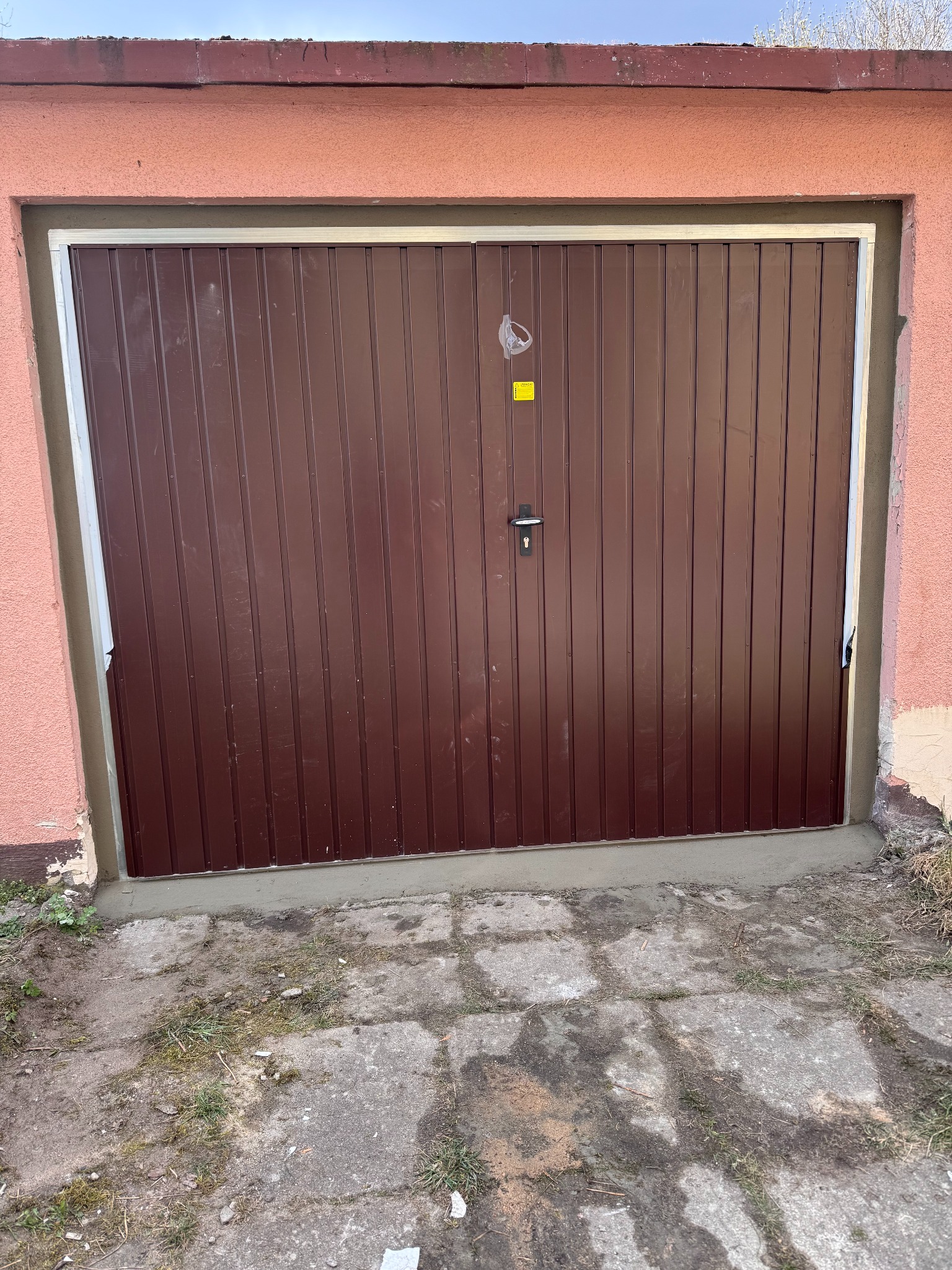 Brązowa, metalowa brama garażowa z pionowymi panelami, świeżo osadzona w ościeżnicy, widoczne ślady cementu i niedoskonałości tynku na ścianie.