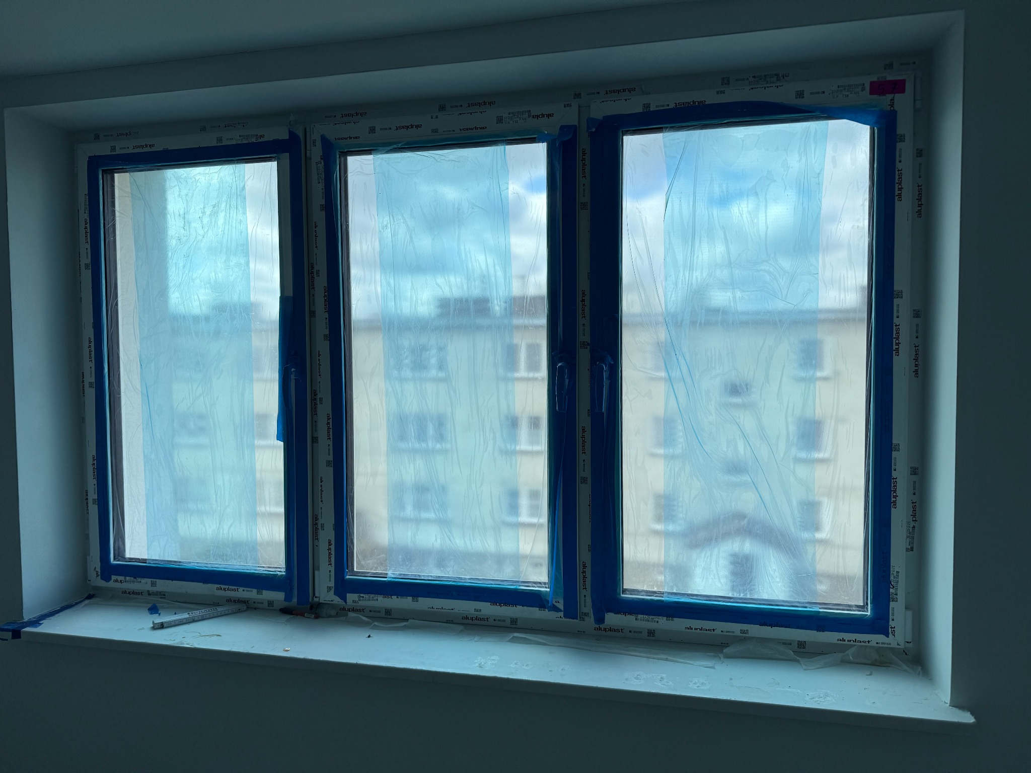 Trzy okna PCV zabezpieczone niebieską folią malarską w trakcie prac wykończeniowych, widoczny budynek za oknem i miarka na parapecie.