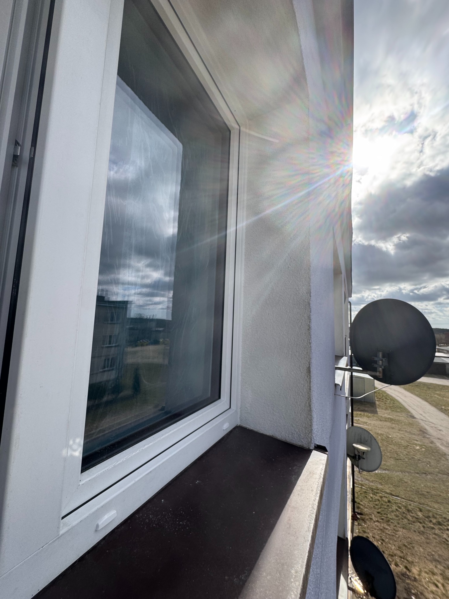 Białe okno z widokiem na krajobraz miejski i anteny satelitarne, oświetlone promieniami słonecznymi.