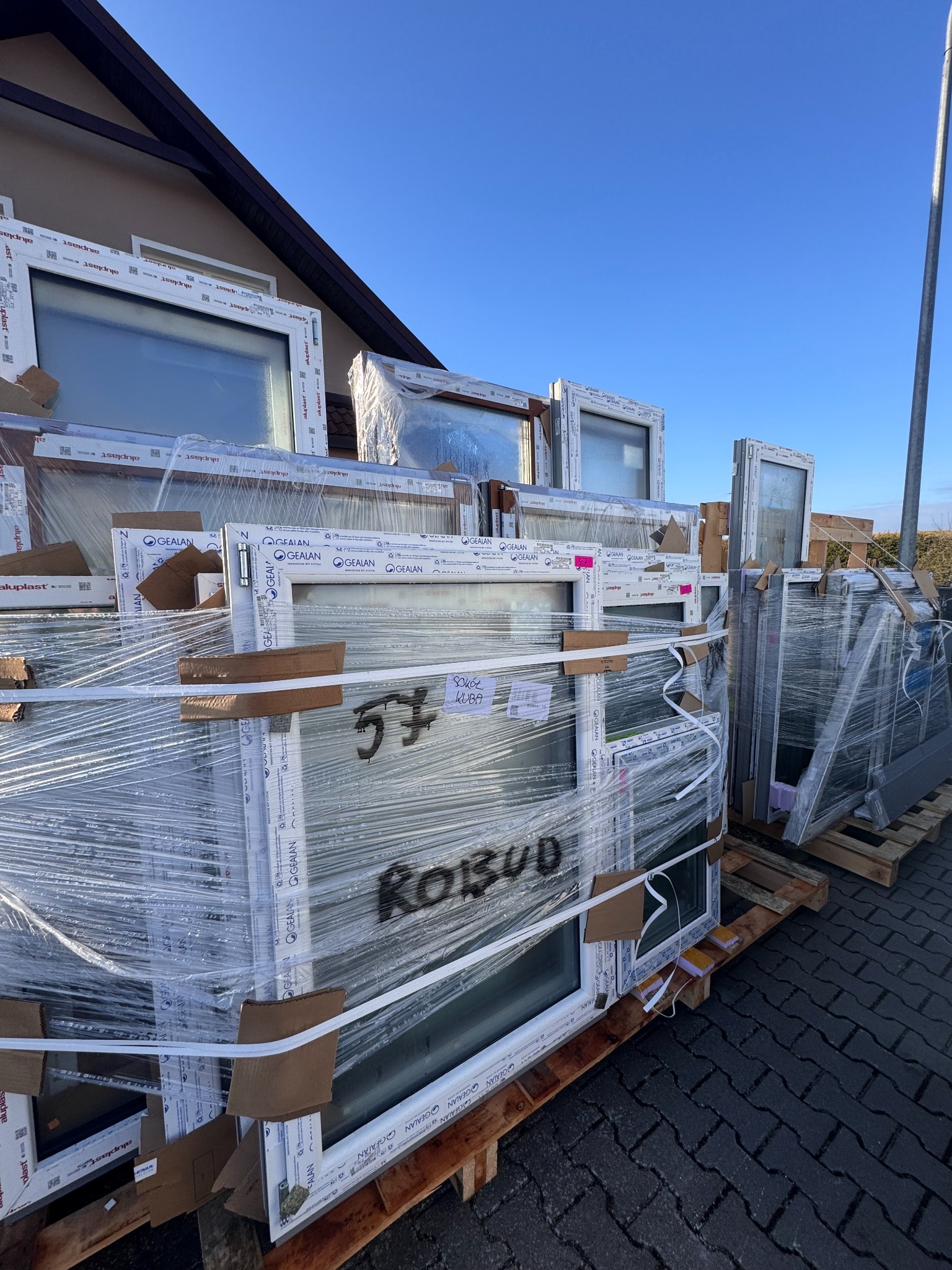 Palety z nowymi oknami PCV, zabezpieczone folią stretch, przygotowane do montażu, widok na budynek w tle i błękitne niebo.