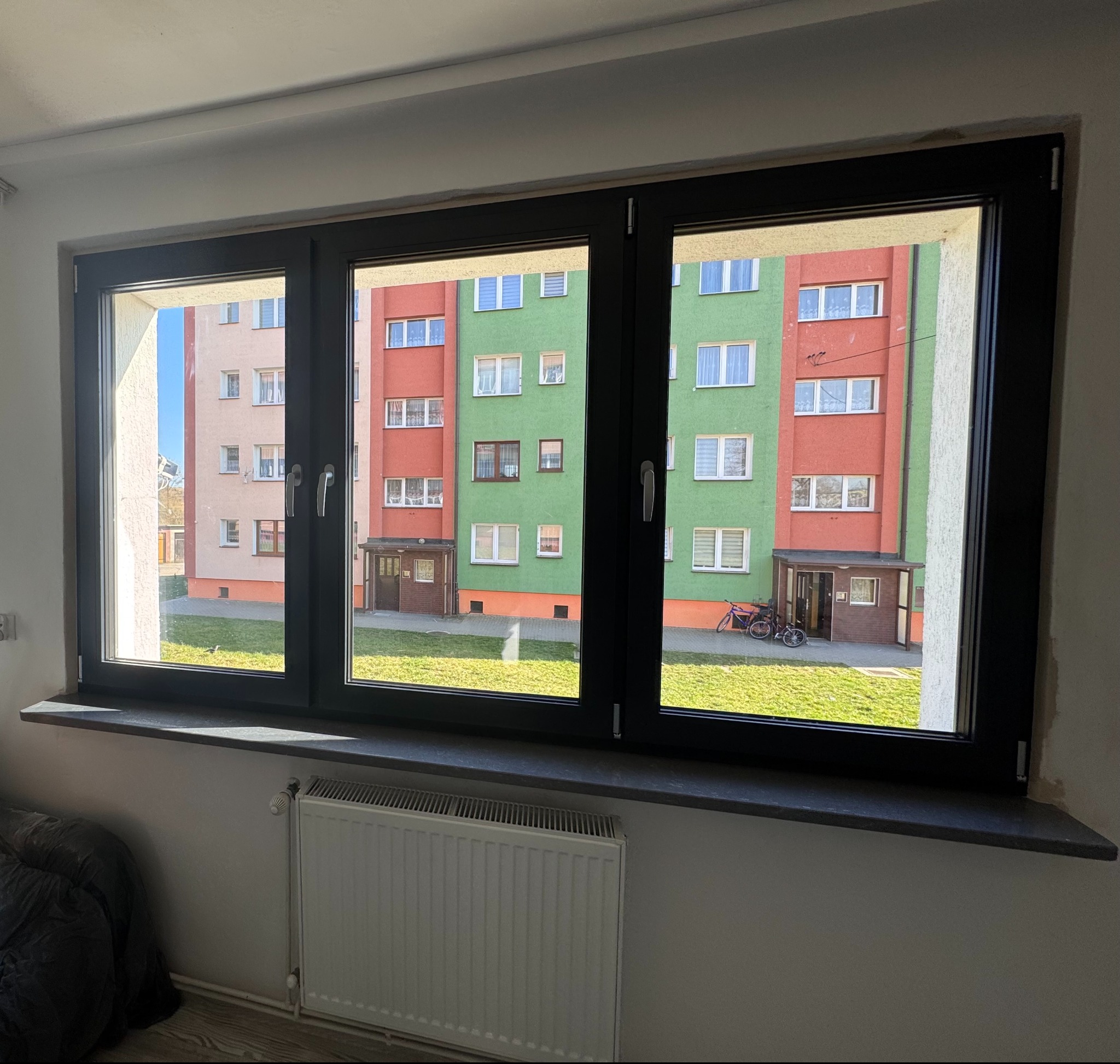 Trzyczęściowe okno z czarną ramą, widok na blok z zielono-czerwonymi elewacjami i trawnik, grzejnik pod parapetem.