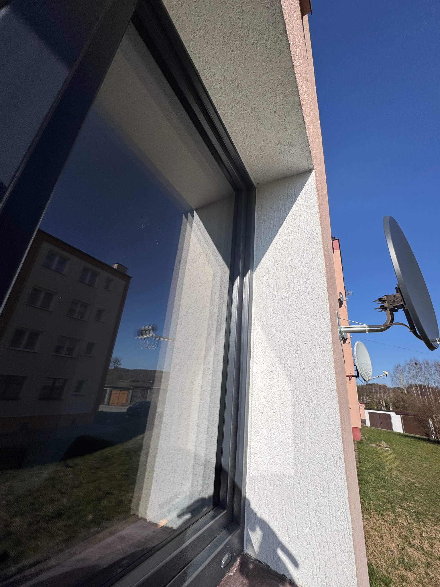 Szare okno z widocznym w szybie odbiciem budynku i nieba, obok elewacja budynku z anteną satelitarną na tle błękitnego nieba.