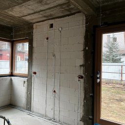 ELLUK TRZECIAK ŁUKASZ - Surowy stan: białe ściany z betonu komórkowego z widocznymi przewodami elektrycznymi w peszlach, puszki instalacyjne w kolorze czerwonym. Okno i drzwi balkonowe.