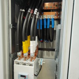 ELLUK TRZECIAK ŁUKASZ - Rozdzielnica elektryczna z grubymi kablami oznaczonymi kolorami, szynami miedzianymi i wyłącznikiem. Widoczne oznaczenia PE i N na szynach. Elementy instalacji wewnątrz szafy.