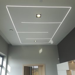 ELLUK TRZECIAK ŁUKASZ - Nowoczesny sufit z oświetleniem LED w kształcie prostokątów i punktowymi lampami. Minimalistyczny design wnętrza z szarą ścianą i jasnymi meblami.