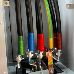 Kable elektryczne w skrzynce rozdzielczej, oznaczone kolorami (zielony, niebieski, czerwony, brązowy) i podłączone do zacisków. Widoczne śruby i metalowe elementy konstrukcyjne.