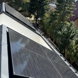 Panele fotowoltaiczne na dachu nowoczesnego domu, widok z góry, otoczenie drzew, słoneczny dzień. Czarny kolor paneli kontrastuje z jasną elewacją budynku.