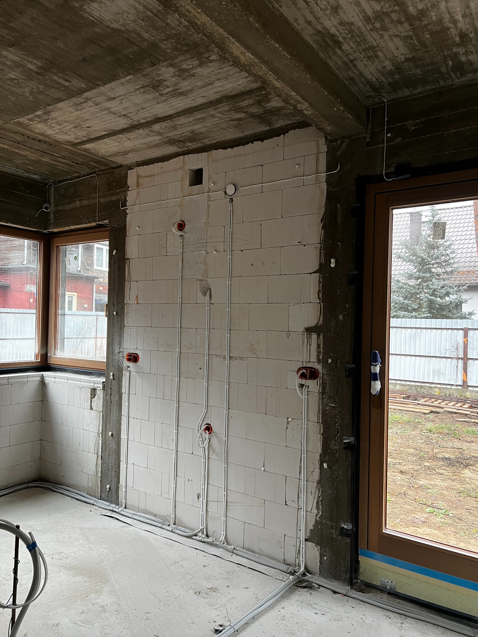 Surowy stan: białe ściany z betonu komórkowego z widocznymi przewodami elektrycznymi w peszlach, puszki instalacyjne w kolorze czerwonym. Okno i drzwi balkonowe.