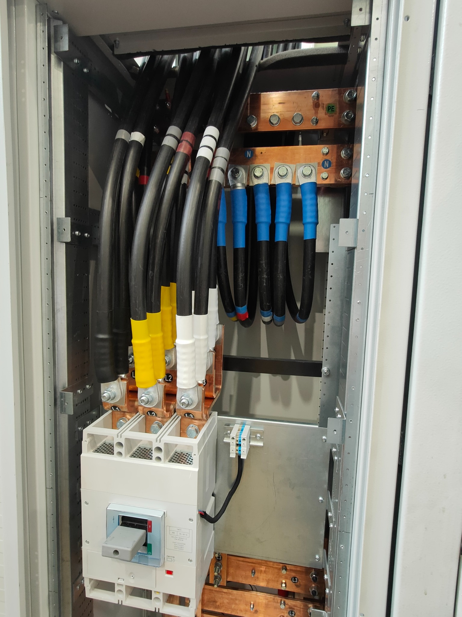 Rozdzielnica elektryczna z grubymi kablami oznaczonymi kolorami, szynami miedzianymi i wyłącznikiem. Widoczne oznaczenia PE i N na szynach. Elementy instalacji wewnątrz szafy.