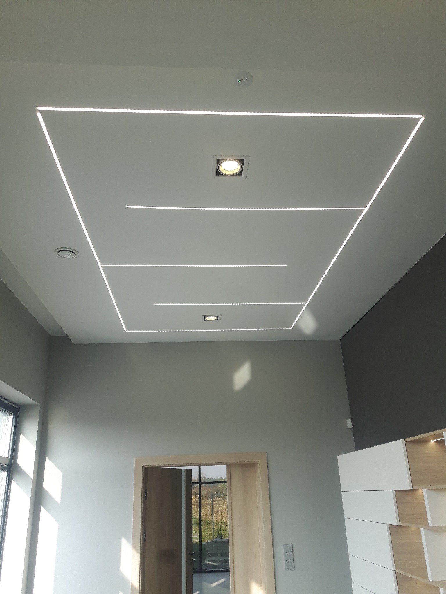 Nowoczesny sufit z oświetleniem LED w kształcie prostokątów i punktowymi lampami. Minimalistyczny design wnętrza z szarą ścianą i jasnymi meblami.