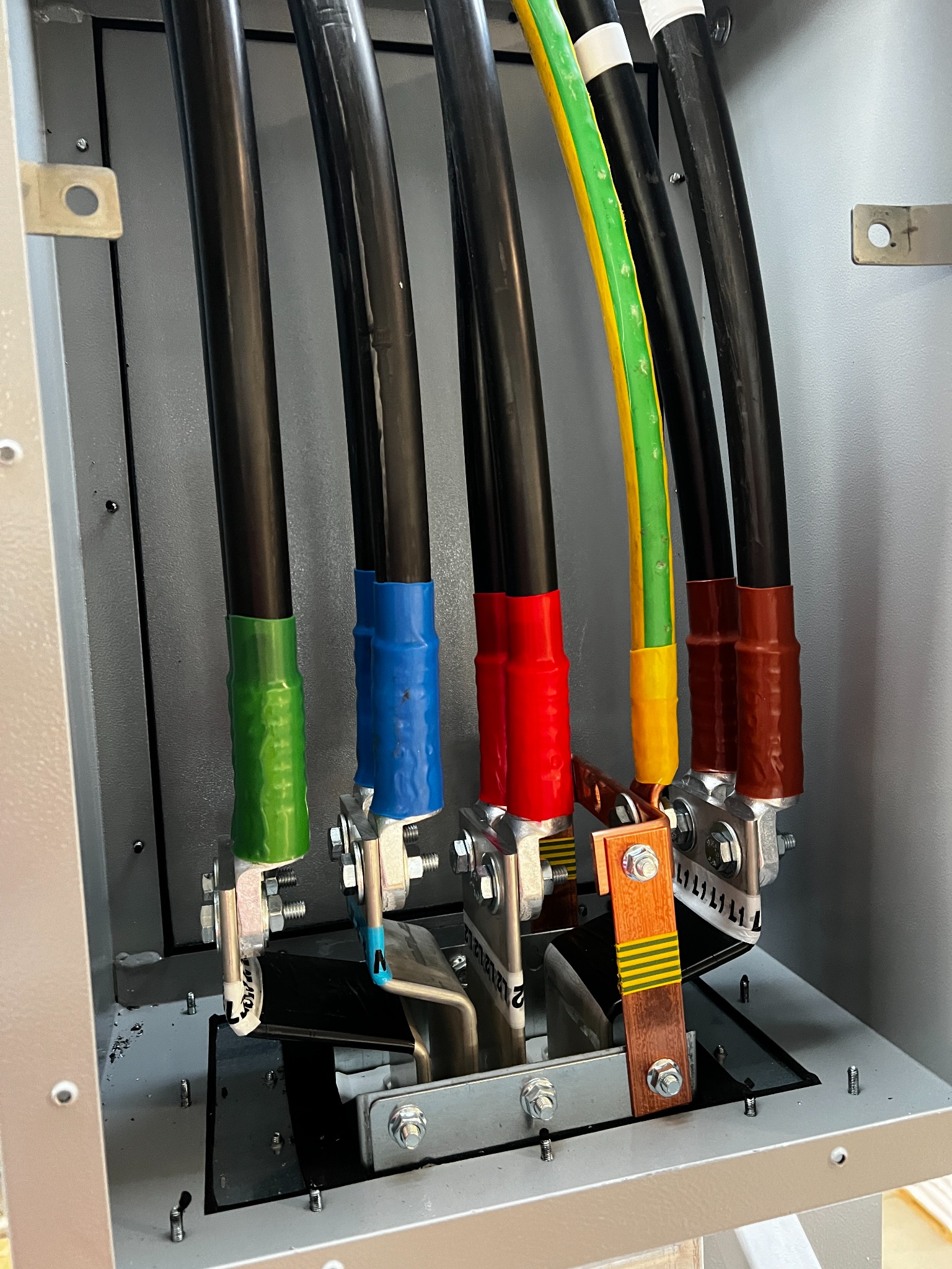 Kable elektryczne w skrzynce rozdzielczej, oznaczone kolorami (zielony, niebieski, czerwony, brązowy) i podłączone do zacisków. Widoczne śruby i metalowe elementy konstrukcyjne.