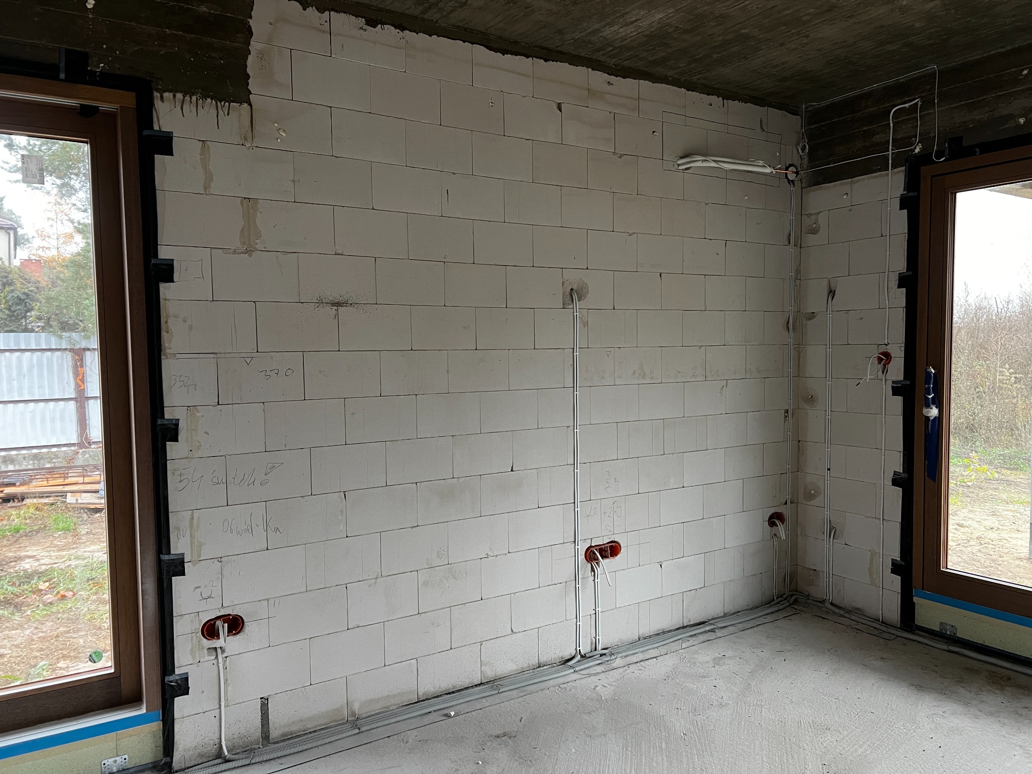 Wewnętrzna instalacja elektryczna w surowym budynku: kable w ścianach z betonu komórkowego, puszki elektryczne, peszle przy podłodze. Widoczne okna i surowy betonowy strop.